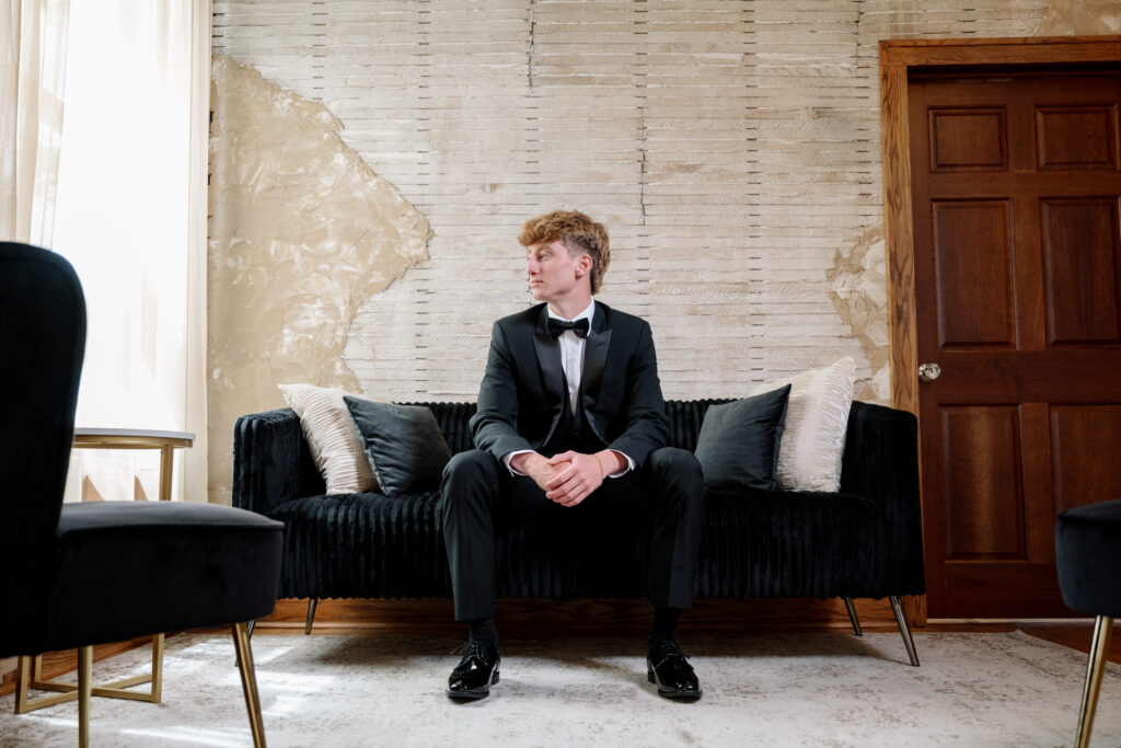 Groom relaxing in a black tuxedo on a couch before the wedding ceremony.