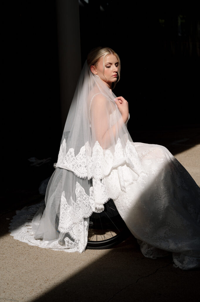 Editorial bridal portrait of a bride in a wheelchair, draped in lace and veil with soft, glowing light