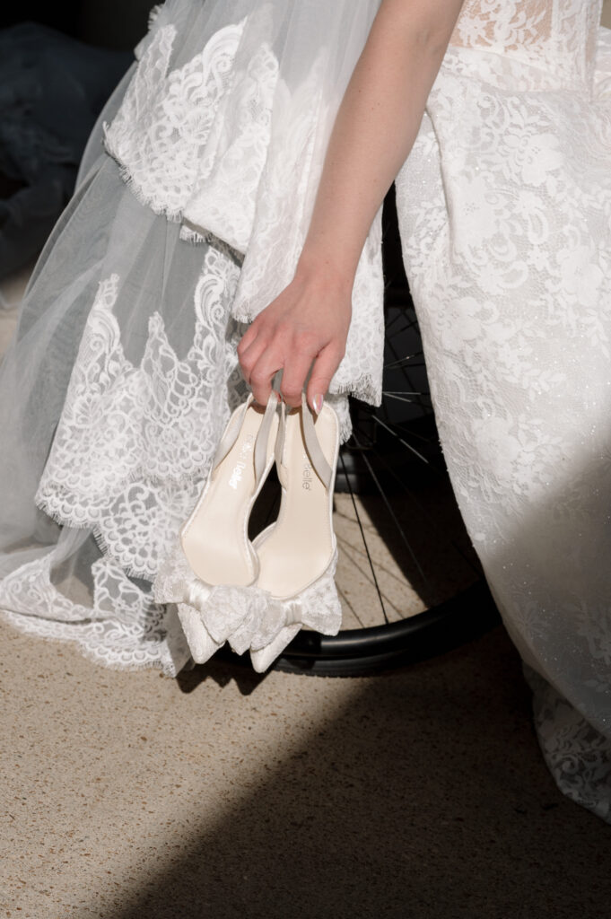 Close-up of bride in lace gown holding wedding shoes next to wheelchair, highlighting inclusive wedding photography